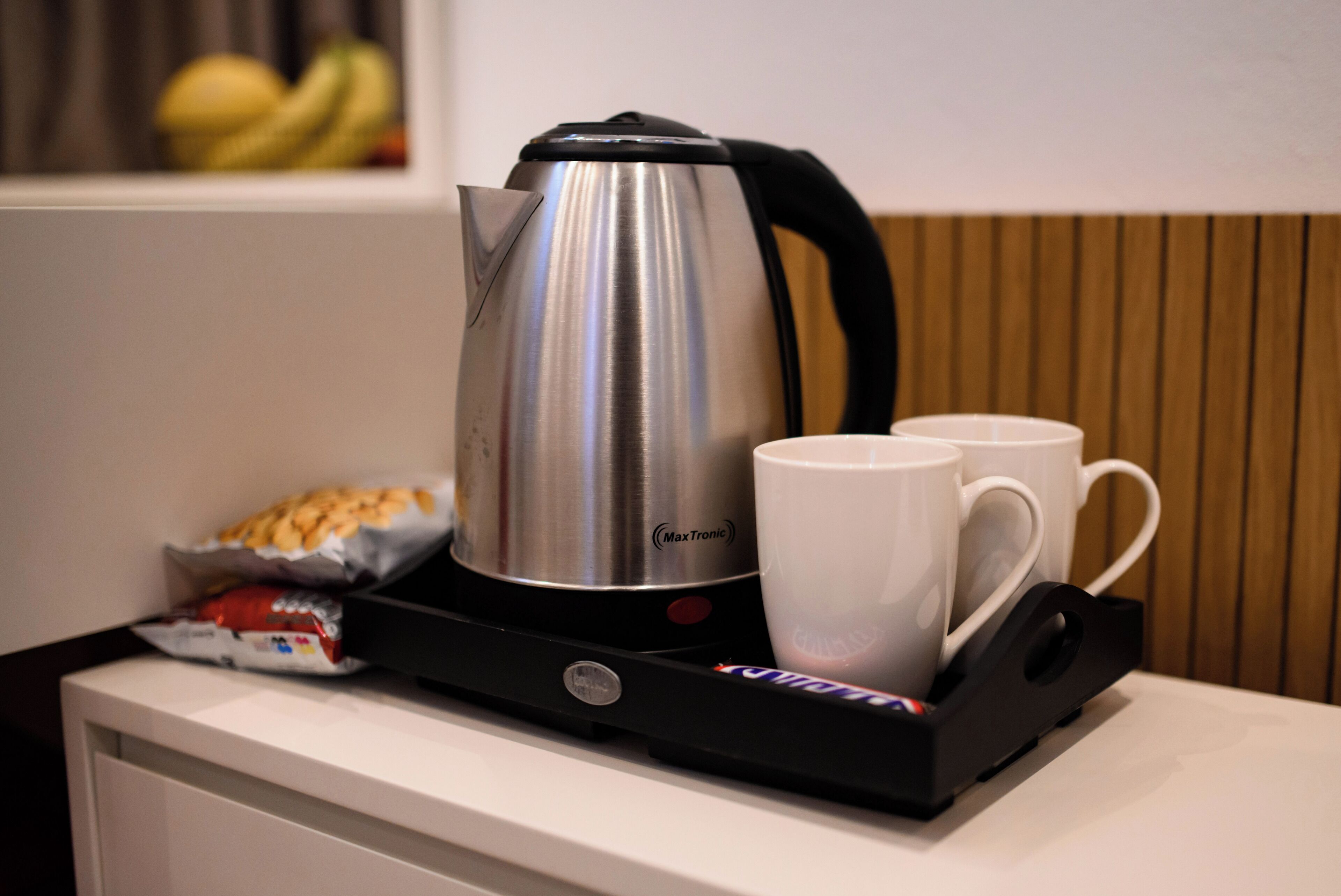 A stainless steel kettle, two white cups, and a packet of snacks on a tray.