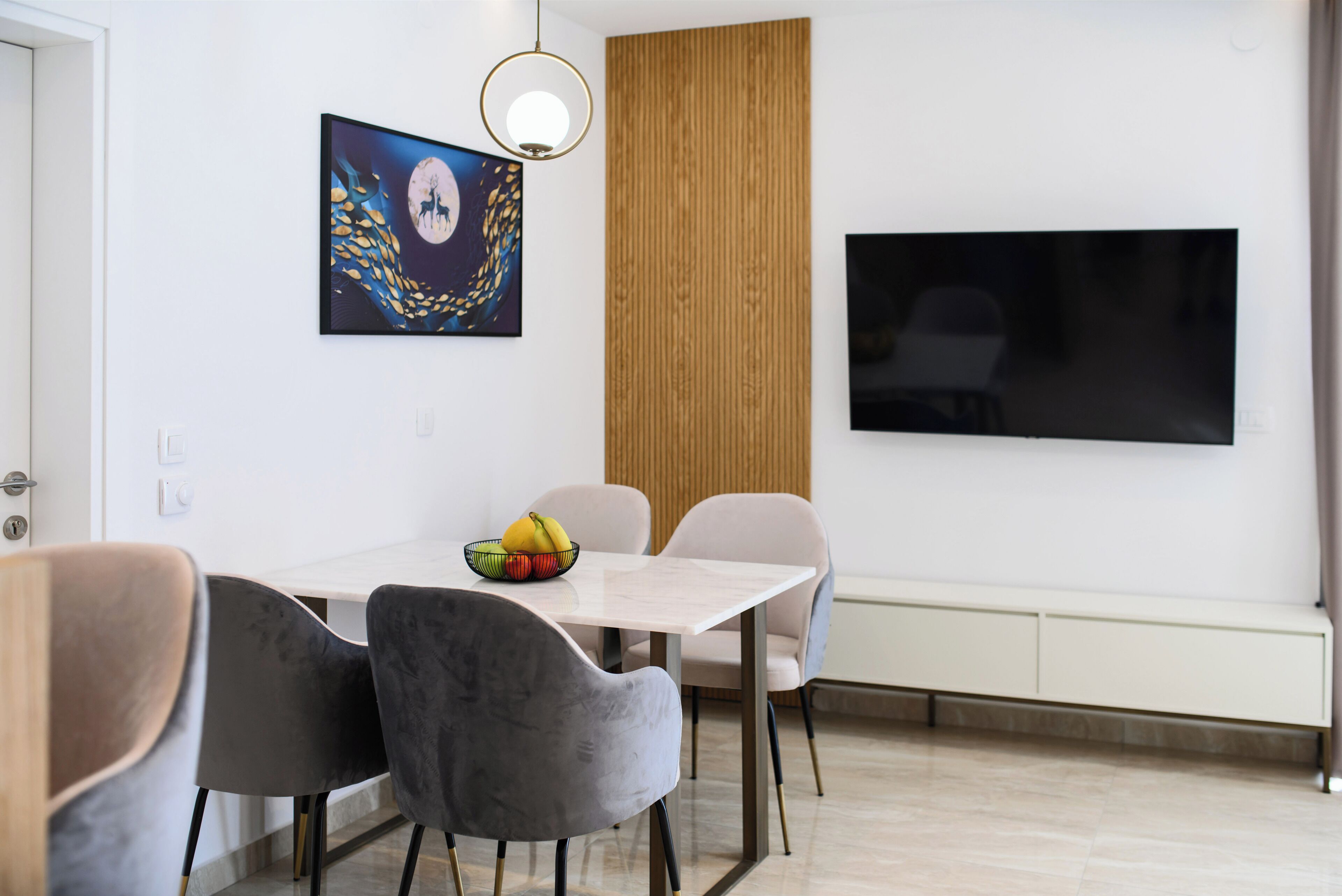 A modern dining area with a white table, grey and beige chairs, a fruit bowl, a wall-mounted painting, and a flat-screen TV.