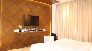 Ein Hotelzimmer mit einem großen Bett, einem flachbildfernseher an einer Holzwand, einem kleinen Schreibtisch mit einer Pflanze und einem Fenster mit Vorhängen.