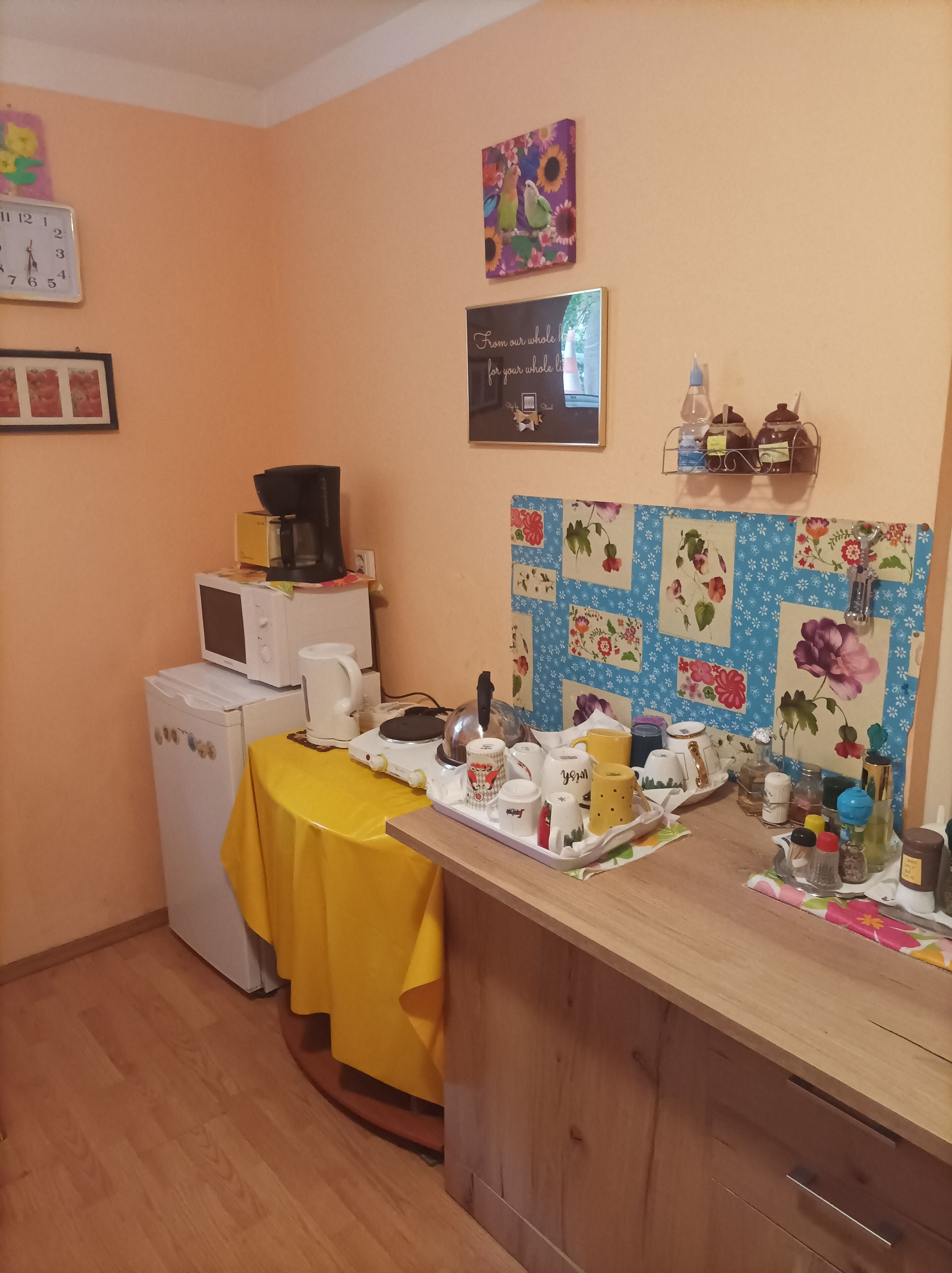 A kitchen with a microwave, kettle, and kettle on a table. There is a clock on the wall and a framed picture above the microwave.