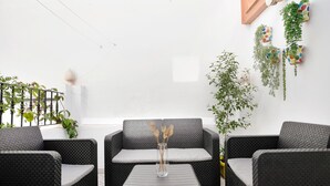 A patio area with wicker furniture, a vase with dried plants, and hanging planters against a white wall.