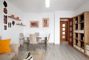 Una sala de estar moderna con una mesa de comedor de vidrio, sillas color beige, un sofá con un cojín naranja, estanterías de madera con objetos decorativos y dos obras de arte enmarcadas en la pared.