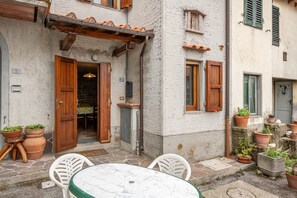 Un patio con una mesa y sillas, plantas en macetas y la entrada de un edificio con contraventanas de madera.