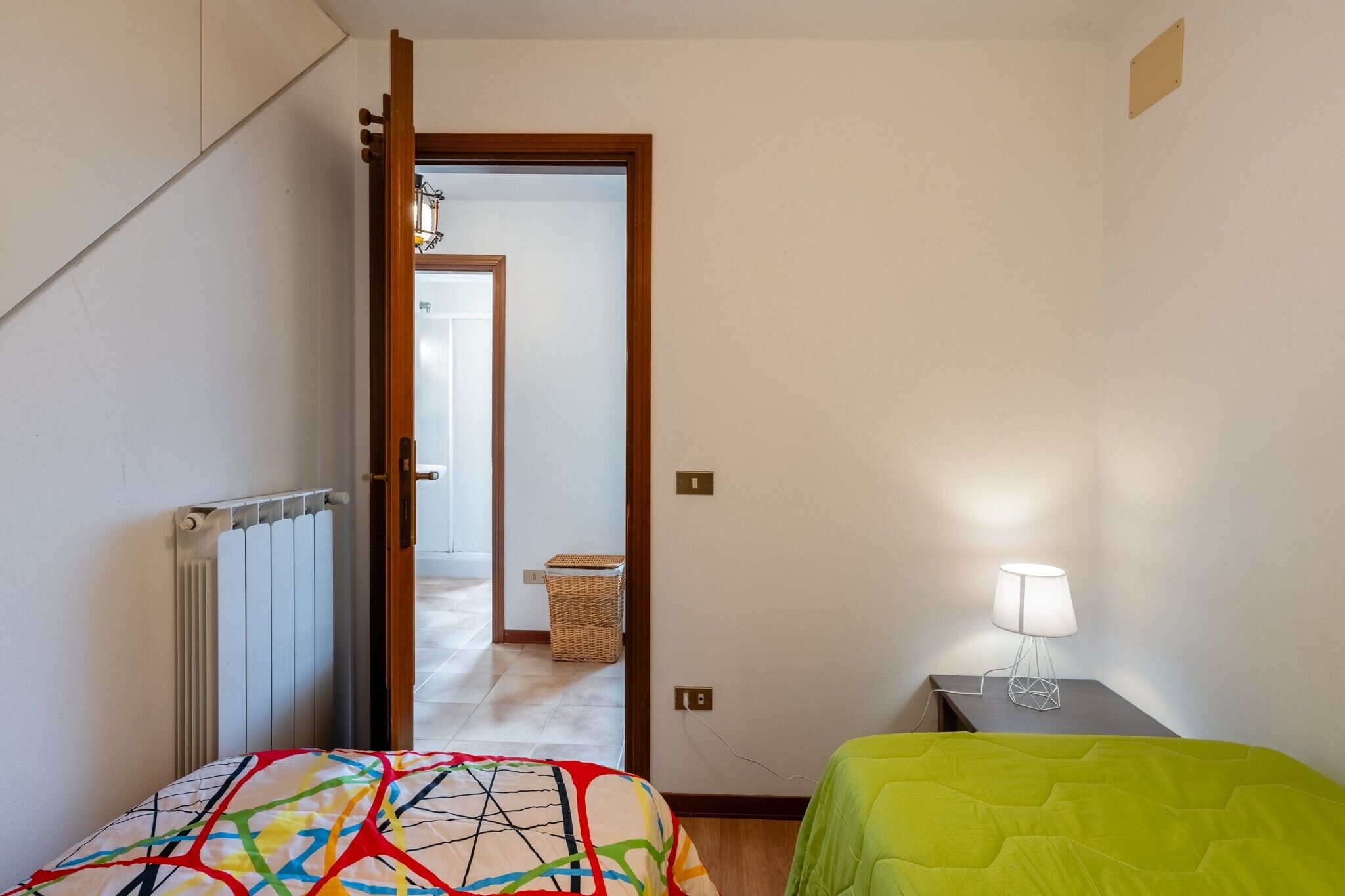 A bedroom with a bed, a bedside table with a lamp, a radiator, a door leading to another room, and a wicker basket.