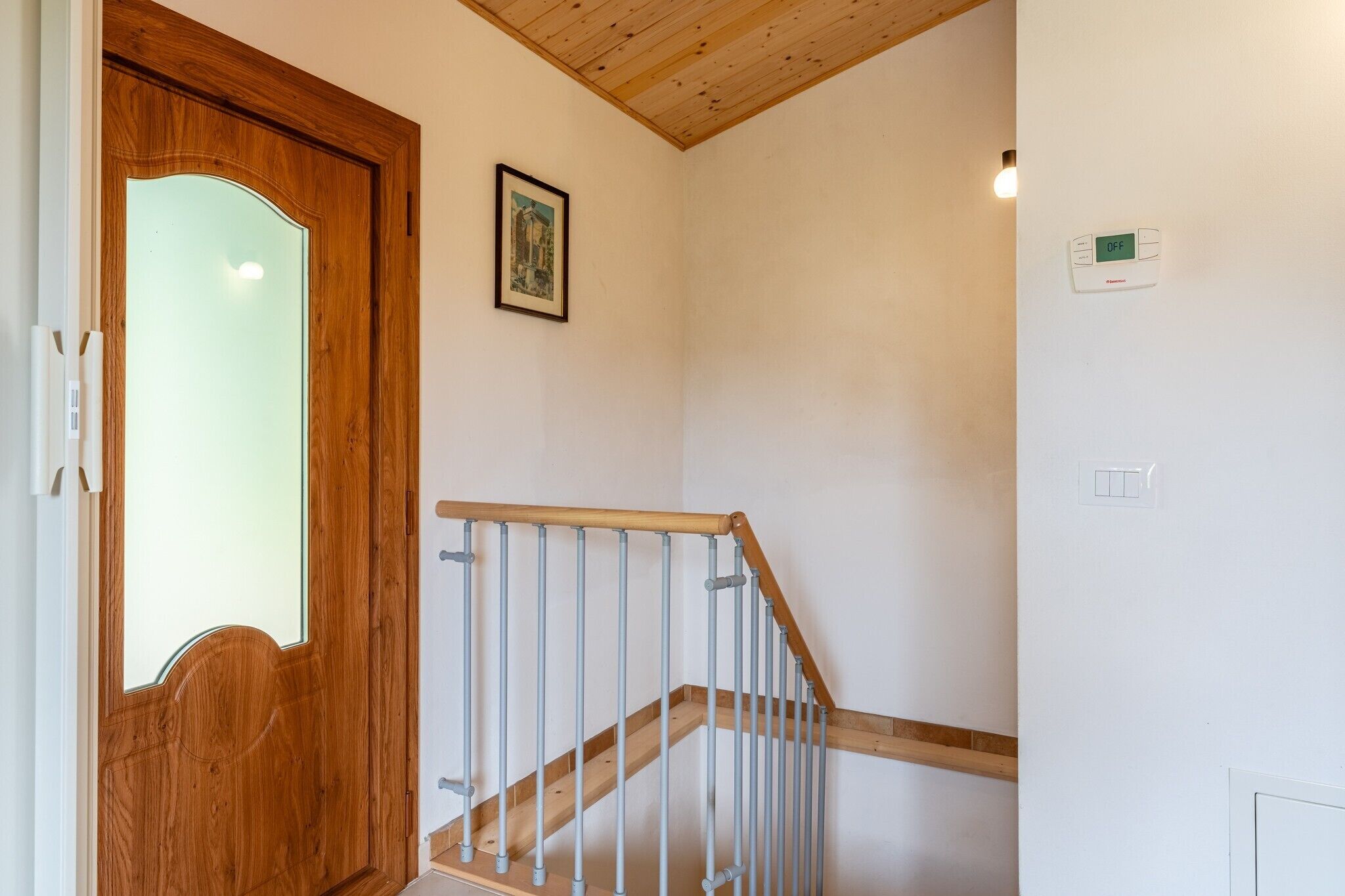A wooden door with a glass panel, a framed picture on the wall, a thermostat, and a light switch.