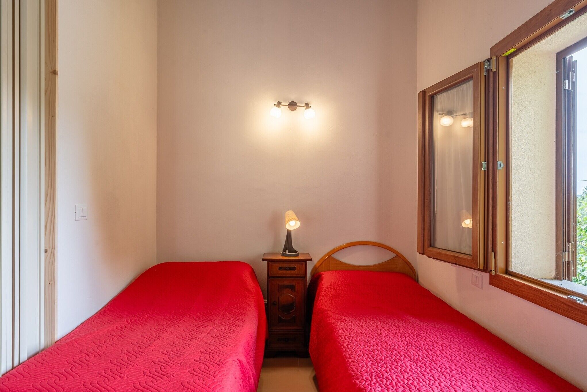 A small room with two single beds, a nightstand with a lamp, a wall-mounted light fixture, and a window with a wooden frame.
