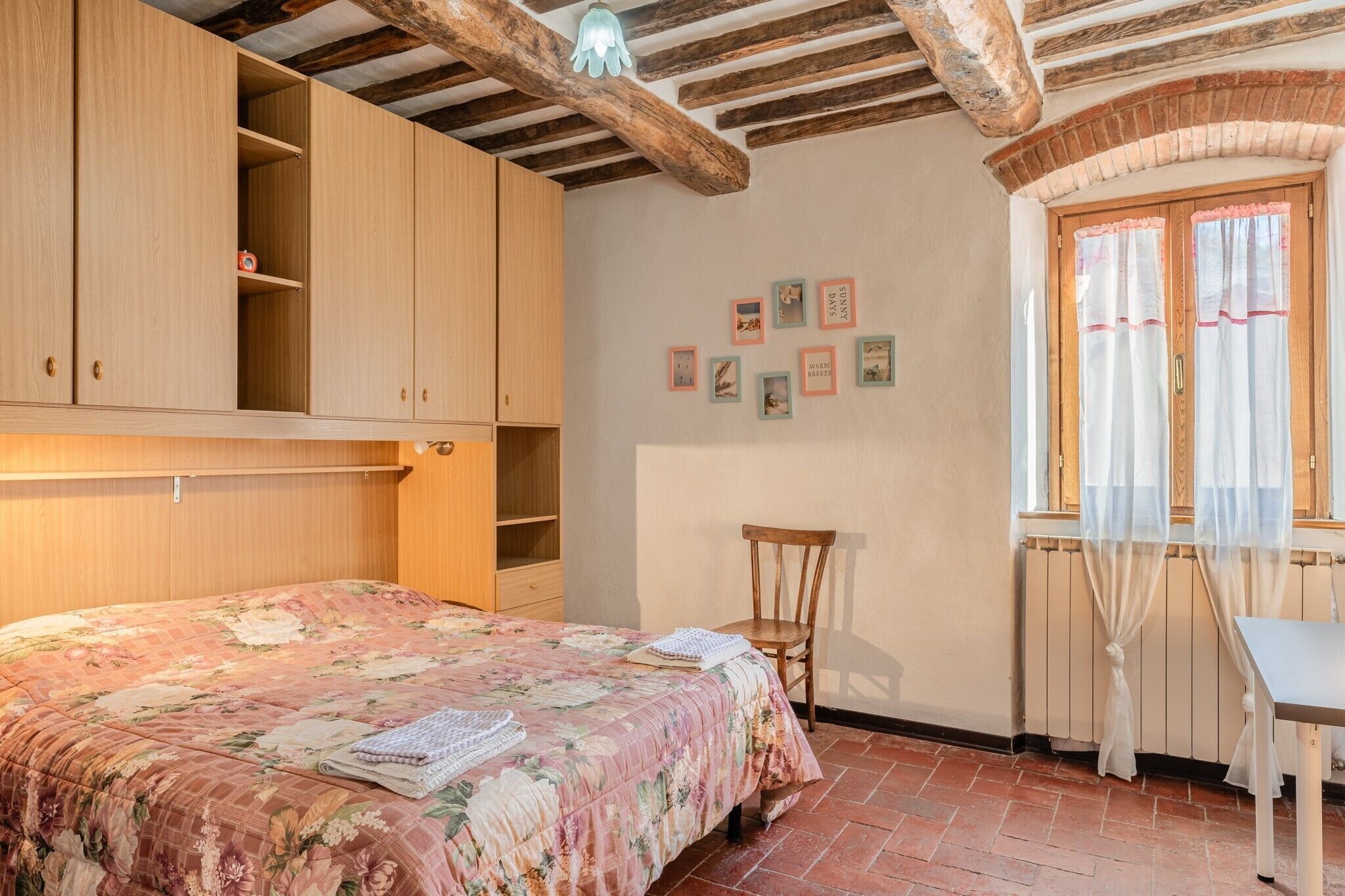 A bedroom with a bed, wooden wardrobe, a chair, a table, and a window with curtains.