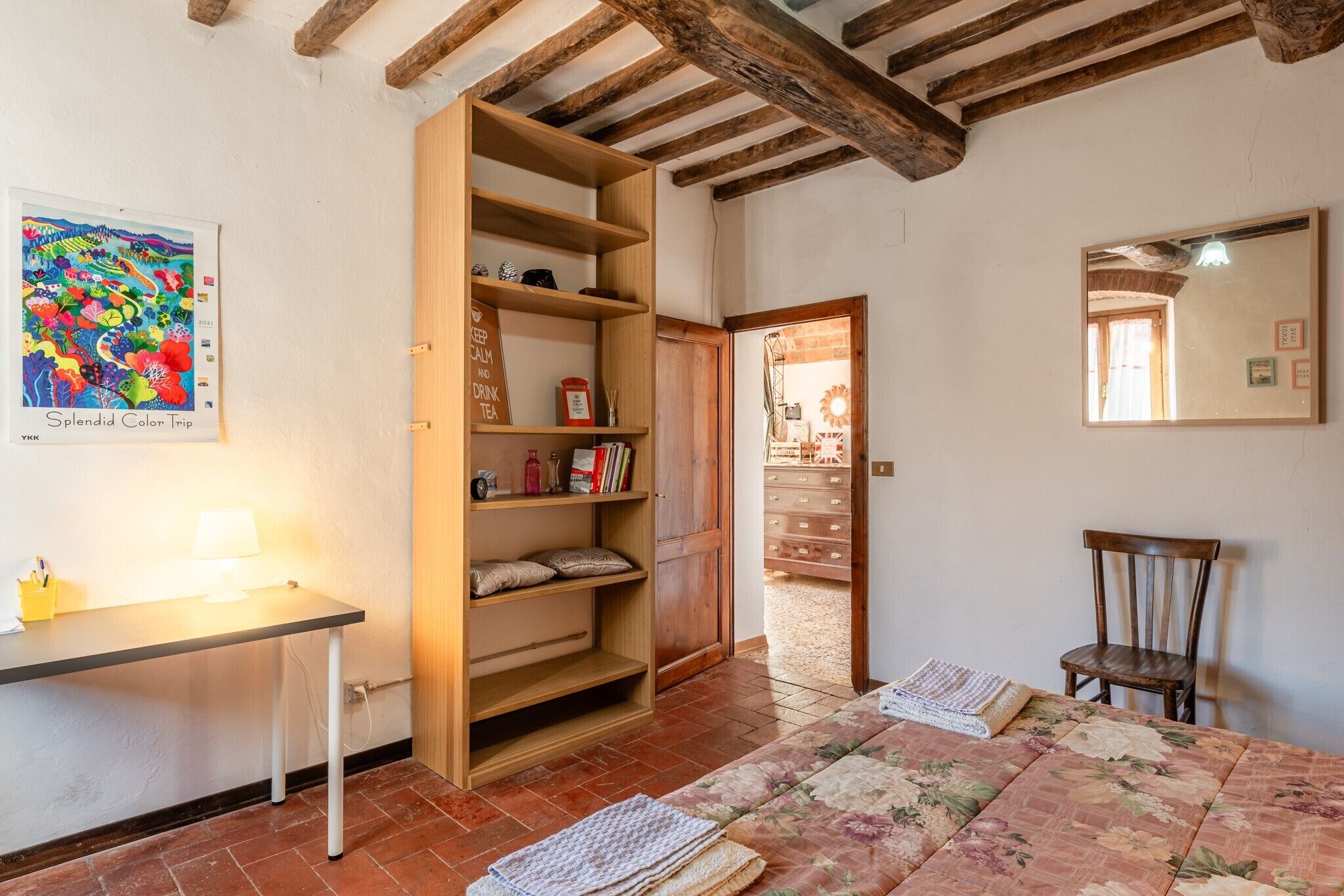 Une pièce avec une bibliothèque en bois, un bureau avec une lampe, un lit recouvert d’une couvre-lit à motifs floraux et un miroir au mur.