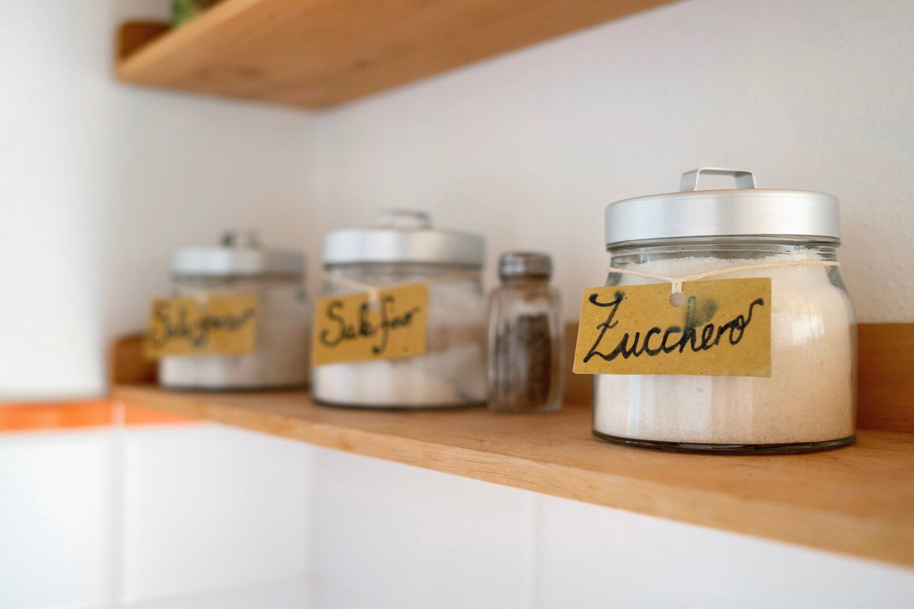 Trois bocaux en verre, dotés de couvercles en métal, sont posés sur une étagère en bois. Chaque bocal porte une étiquette avec un texte écrit à la main.
