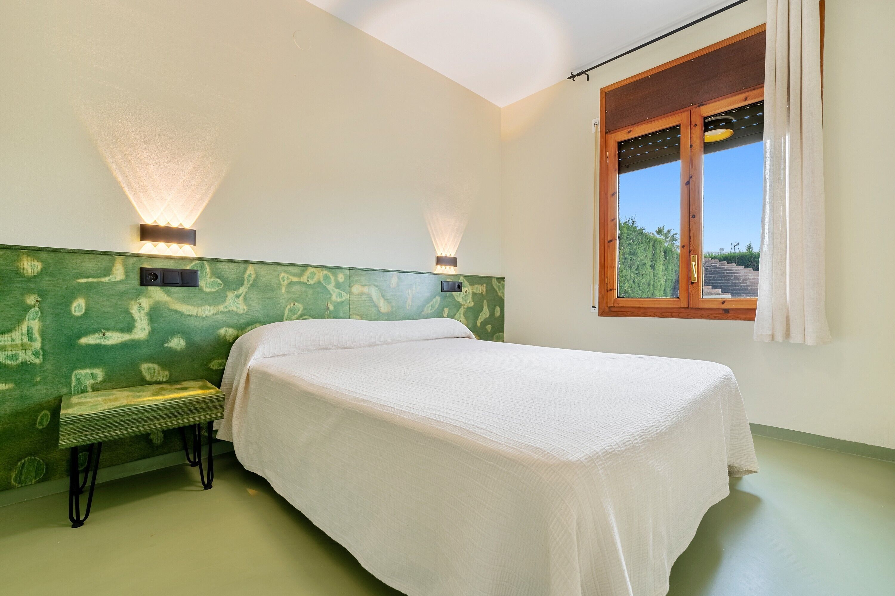 Una habitación con cama, banco, luces fijadas a la pared y ventana con vistas.