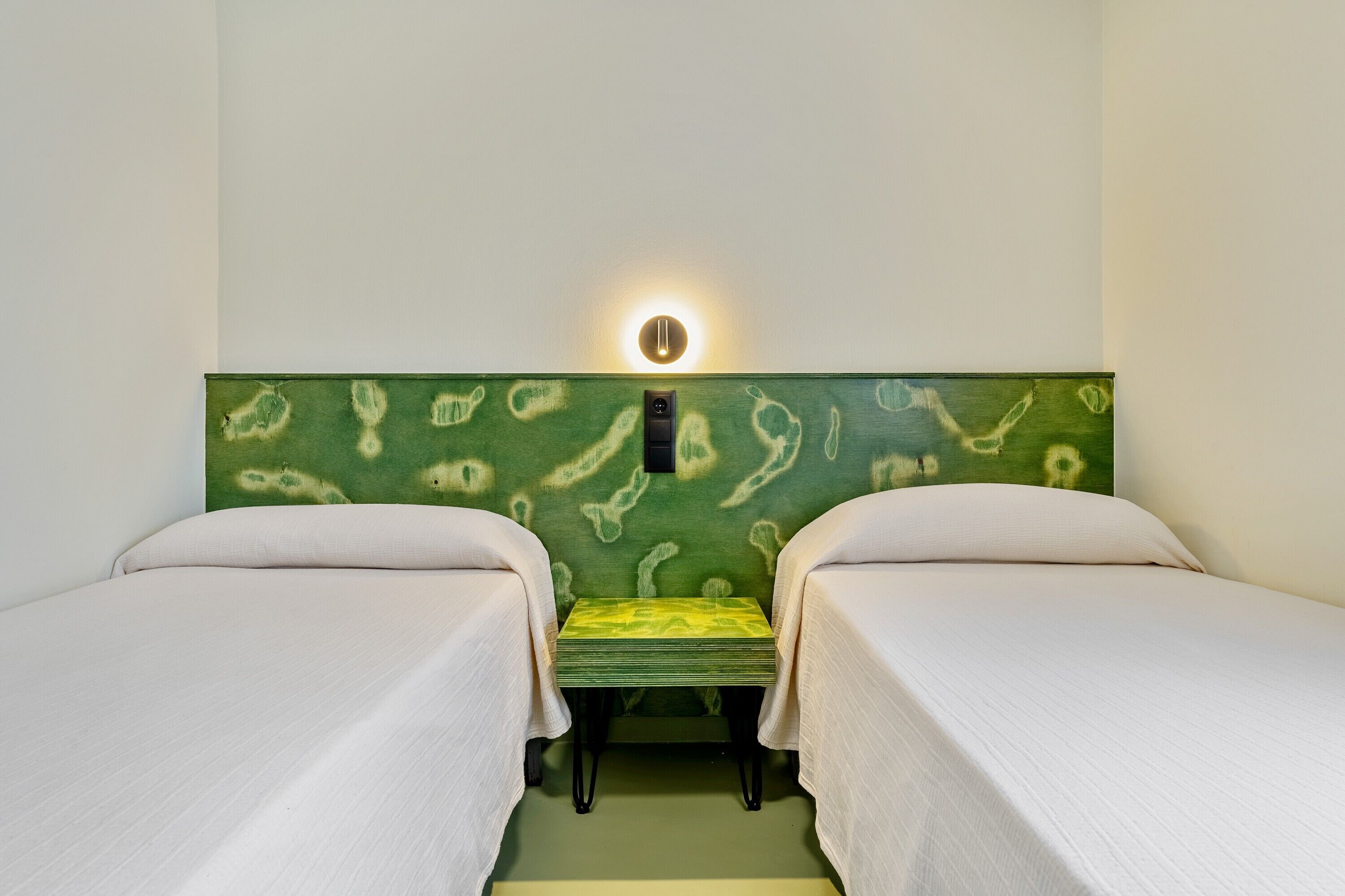 A room with two beds, a green headboard, a small table, and a wall-mounted light.