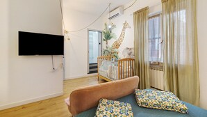 Ein Wohnzimmer mit an der Wand montiertem Fernseher, einem Babybett, einer Couch mit gemusterten Kissen und einem Giraffenwandbild.
