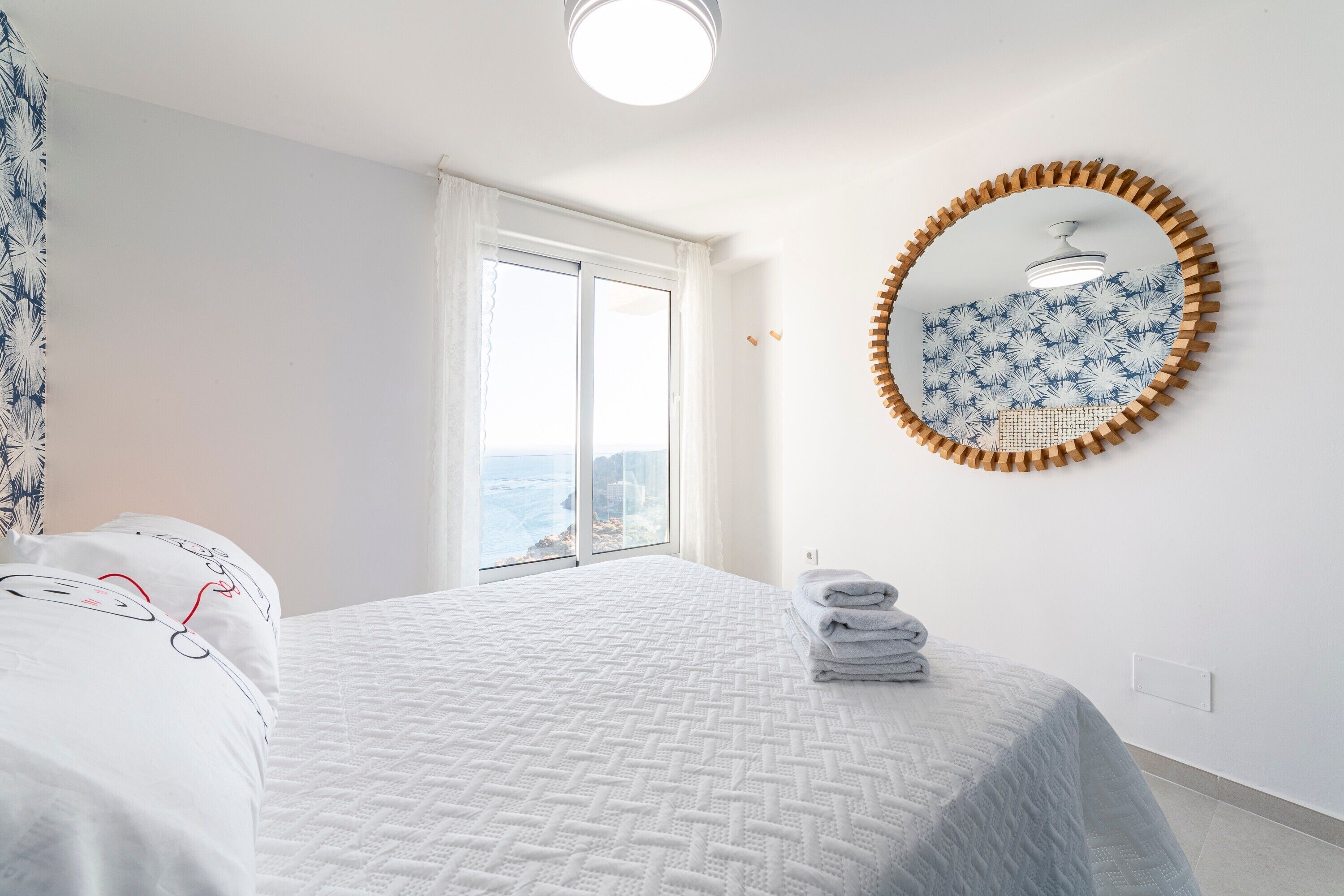 A bedroom with a bed, a window with curtains, a round mirror, and a view of the sea.