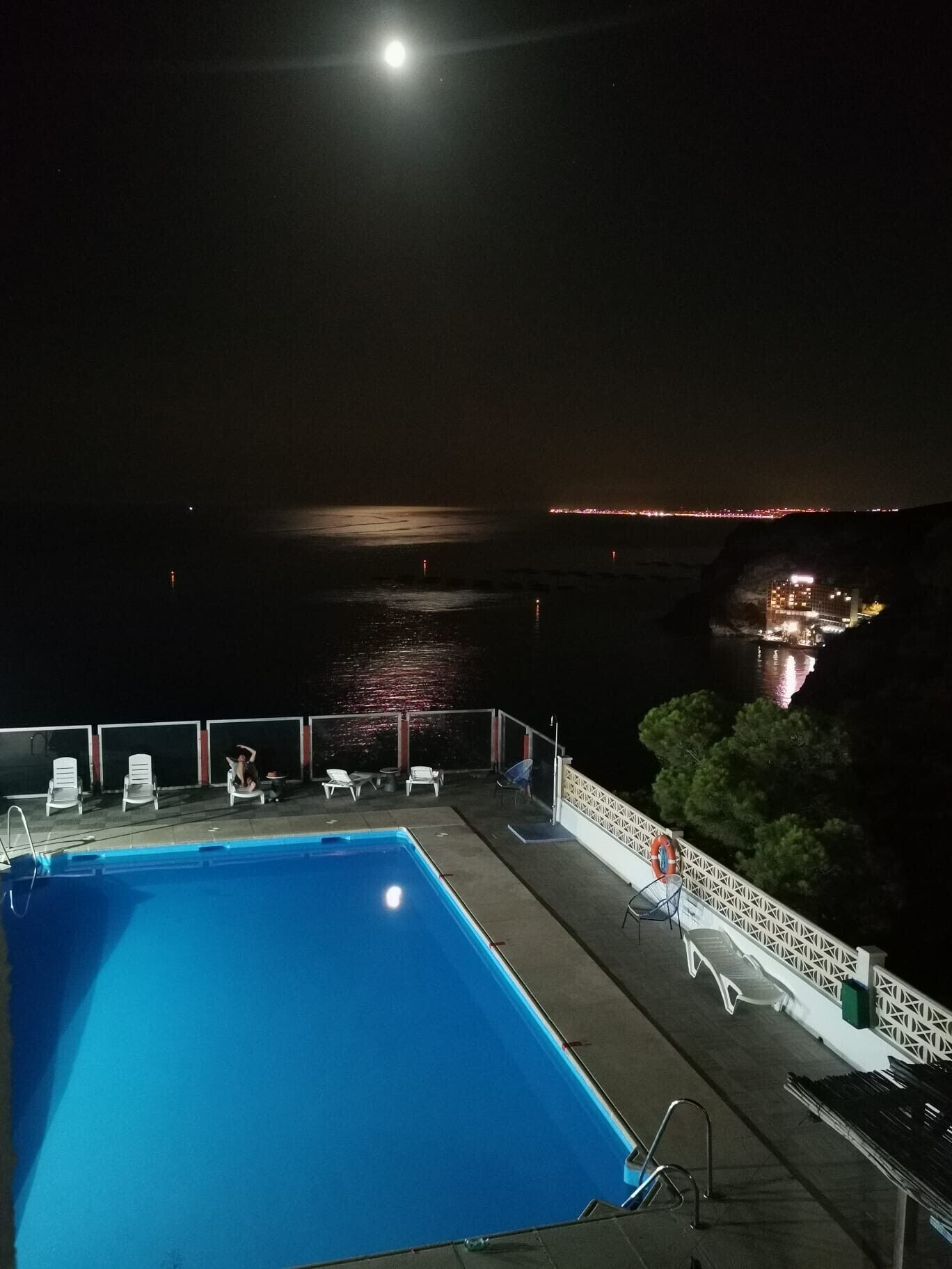 Vista noturna de uma piscina com espreguiçadeiras e corrimão, com vista para um corpo d'água e luzes da cidade ao longe.