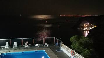 Vista noturna de uma piscina com espreguiçadeiras e corrimão, com vista para um corpo d'água e luzes da cidade ao longe.