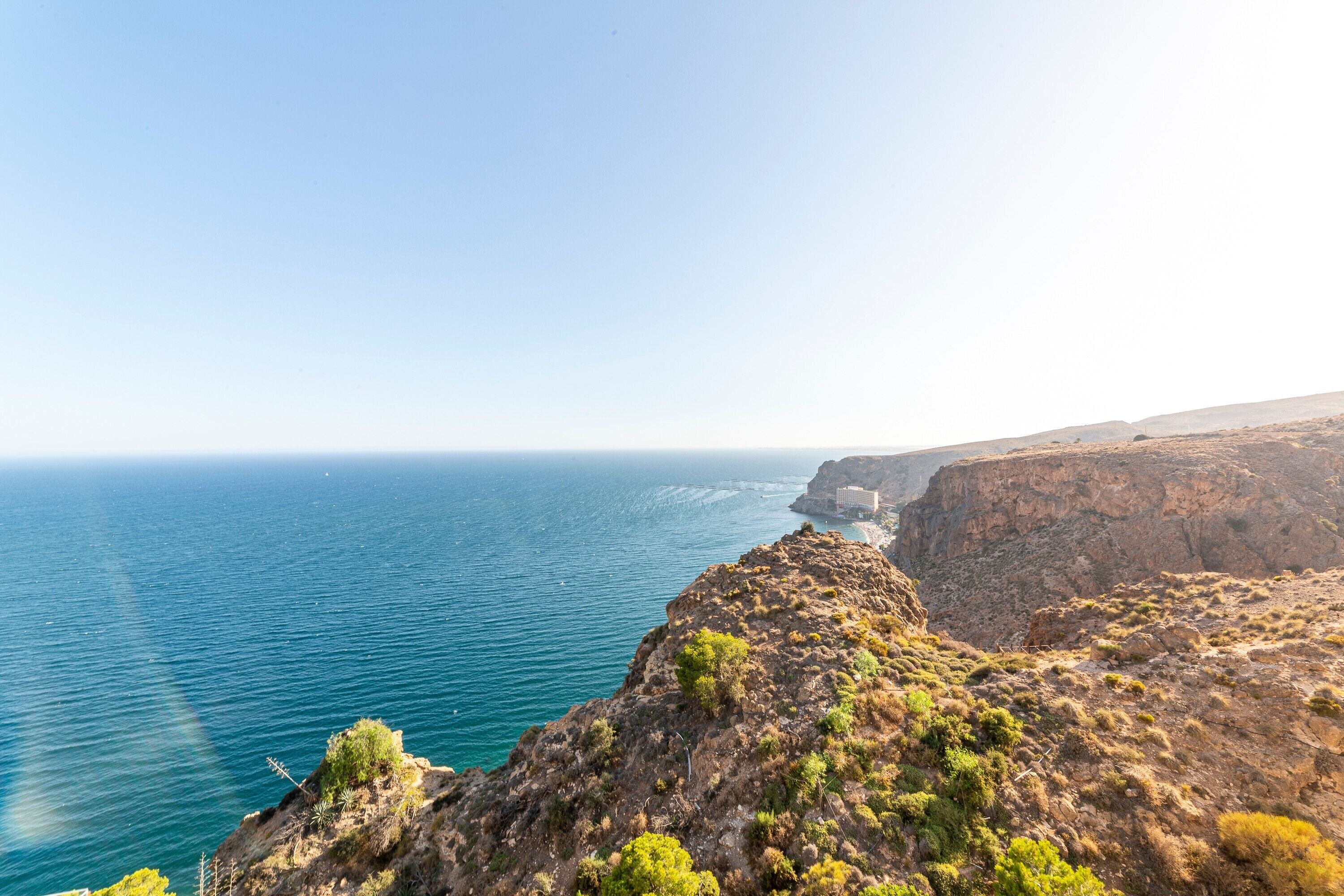 A coastal landscape with a cliff overlooking the sea, a clear sky, and distant buildings.