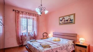 Een slaapkamer met een bed, nachtkastje, stoel, raam met gordijnen, een aan de muur hangend schilderij en een plafondlamp.