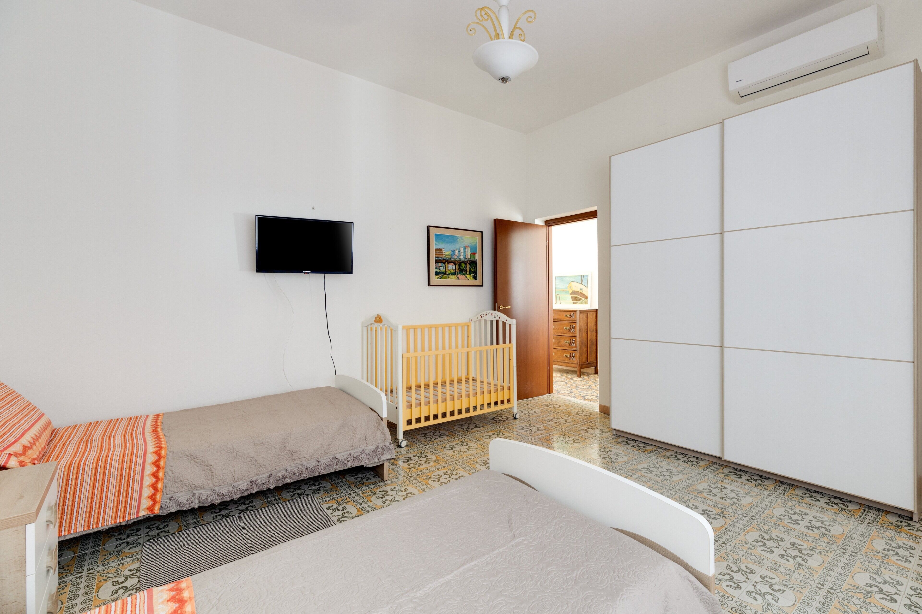 Un dormitorio con cama, cuna, armario, televisor y puerta que da a un balcón.