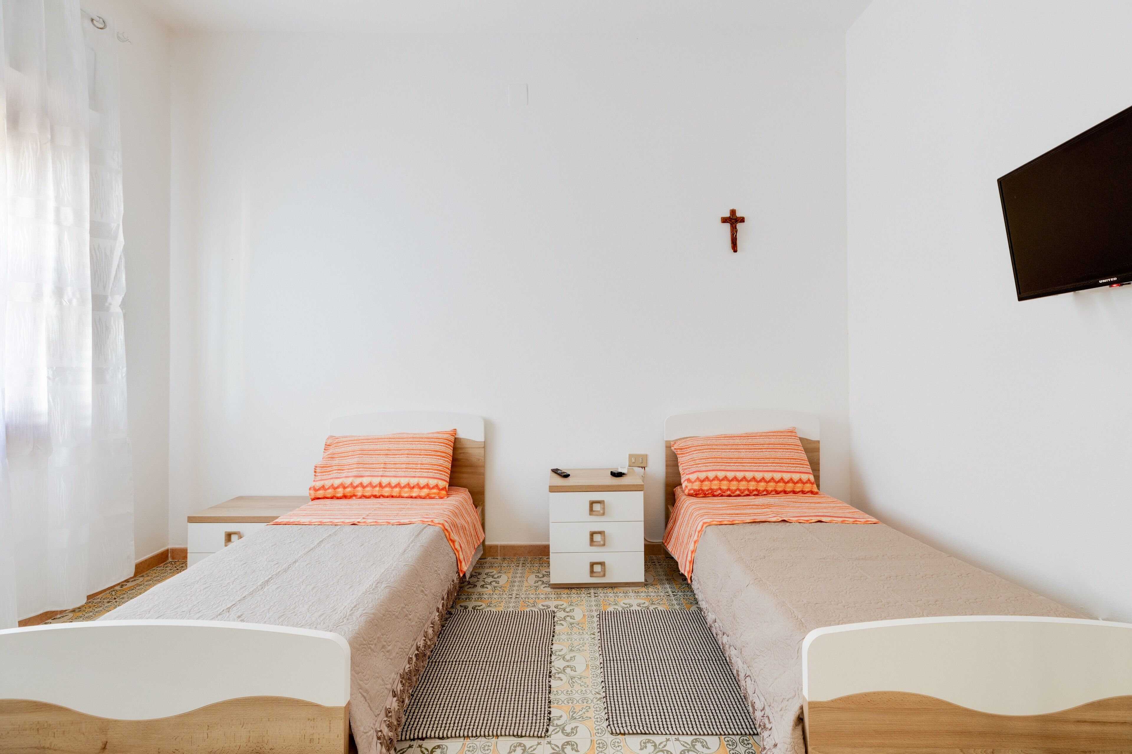 Ein Doppelzimmer mit zwei Betten, einem kleinen Nachttisch, einem an der Wand befestigten Fernseher und einem Kreuz.