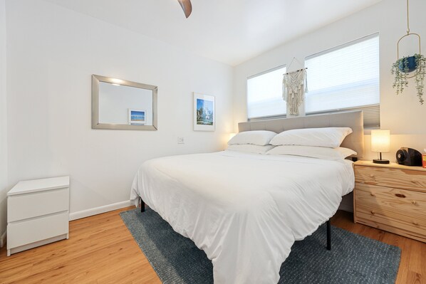 A modern bedroom with a large bed, a bedside table with a lamp, a small white dresser, and a window with a view.