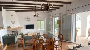 A spacious living room with a fireplace, a dining area with wooden chairs, a sofa, and a TV mounted on the wall. - Villa 'Casa Hyam' with Lake View, Private Pool and Air Conditioning (Alcaucin)