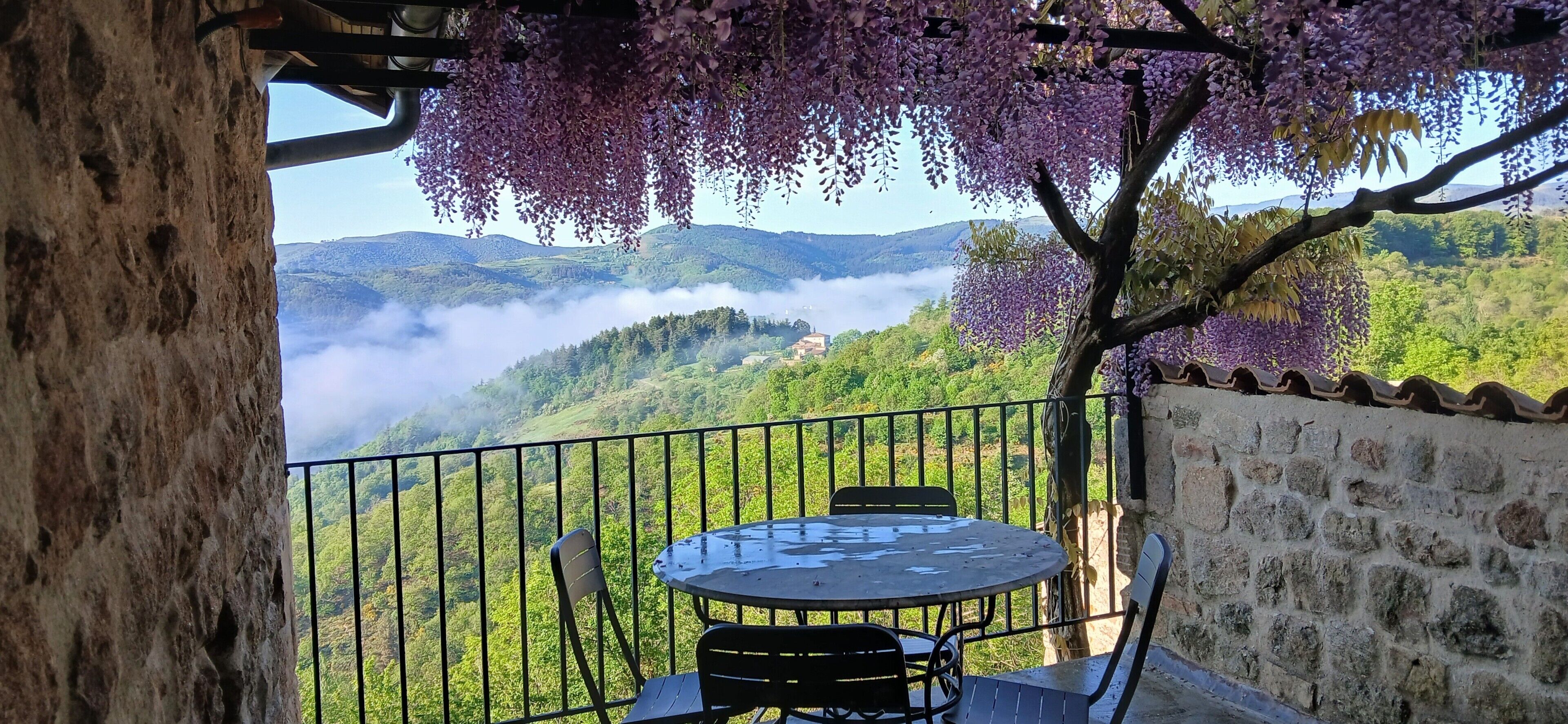 A balcony with a table and chairs, overlooking a mountainous landscape with greenery and a view of the sky.