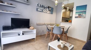 A modern living room with a flat-screen TV, a dining table with chairs, a sofa, a coffee table, and a kitchen area with a sink and cabinets.