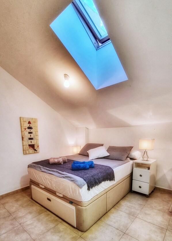 A bedroom with a skylight, a bed with pillows and a blanket, a bedside table with a lamp, and a wall-mounted artwork.