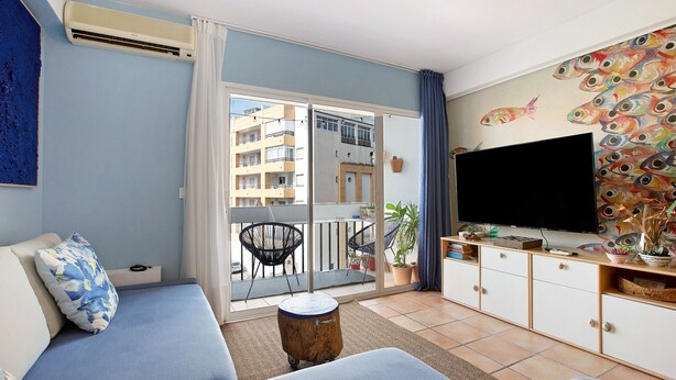 A living room with a sofa, a TV, a balcony with a view of buildings, and a fish mural. - Holiday Apartment "Blue Fish" with Shared Pool, Private Terrace & Wi-Fi (Benalmádena)