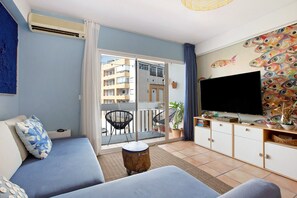 Una sala de estar con sofá, televisor, balcón y vista a edificios, además de un mural de peces.