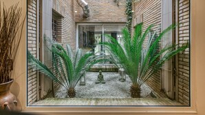 Vista a través de una ventana que muestra un cuarto con pared de ladrillo, una persona sentada y plantas en macetas.