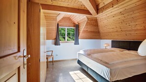 Un dormitorio con techo de madera, ventana con cortinas, cama, una silla y una mesita de noche.