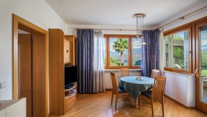 Une pièce comprenant une table à manger, des chaises, un téléviseur et offrant une vue sur la verdure grâce à de grandes fenêtres.