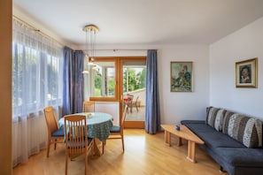 A room with a dining table, chairs, a sofa, a bench, and a view through a glass door.