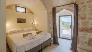 A bedroom with a stone wall, a bed with white linens, a bedside table with a lamp, and a view through a door. - Villa 'Trulli Meridiem Con Piscina' with Private Pool, Wi-Fi and Air Conditioning (Ostuni)