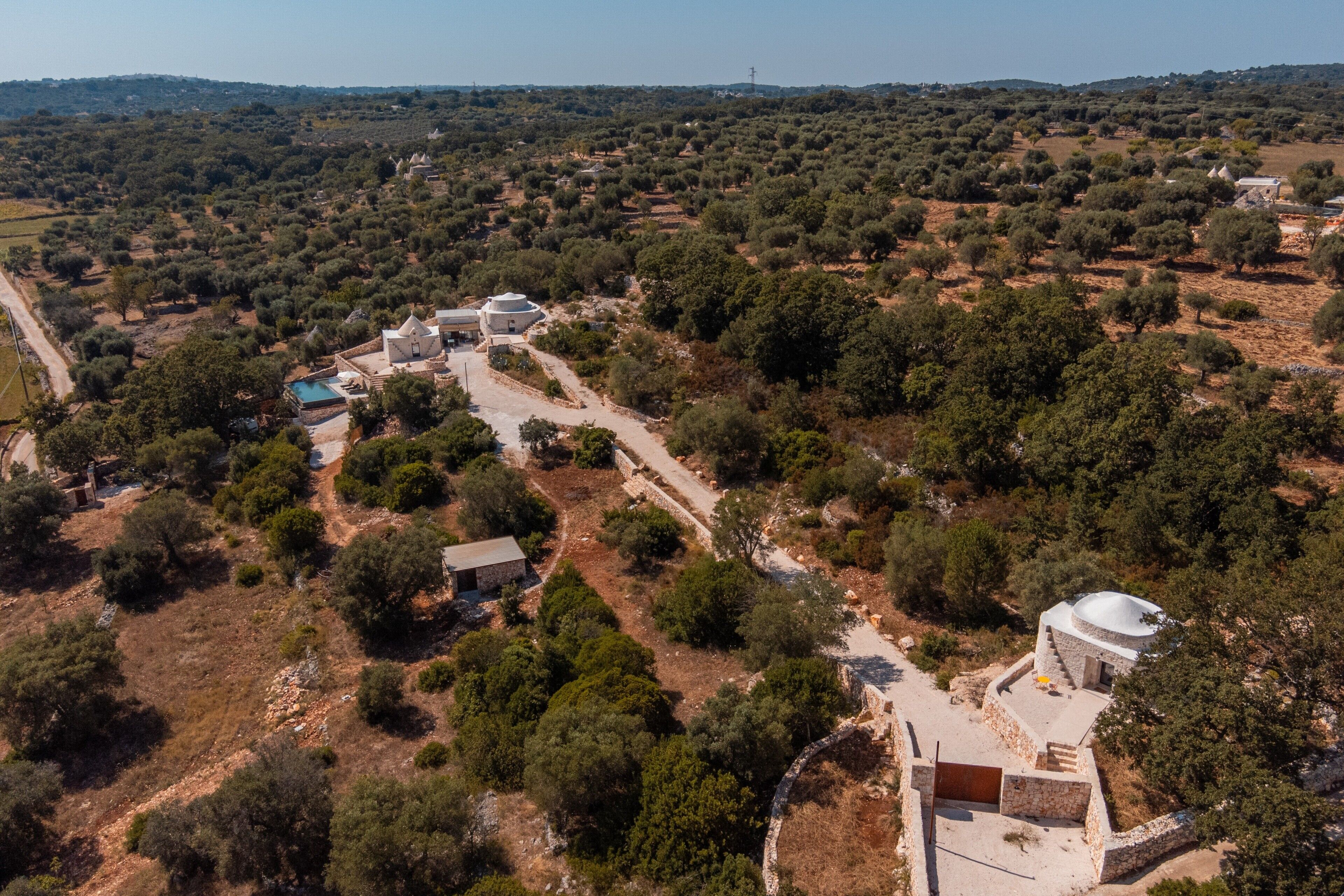 Vista aérea de una zona rural con varias construcciones, incluyendo una gran estructura en forma de cúpula y edificios más pequeños, rodeado de olivos y campos abiertos.