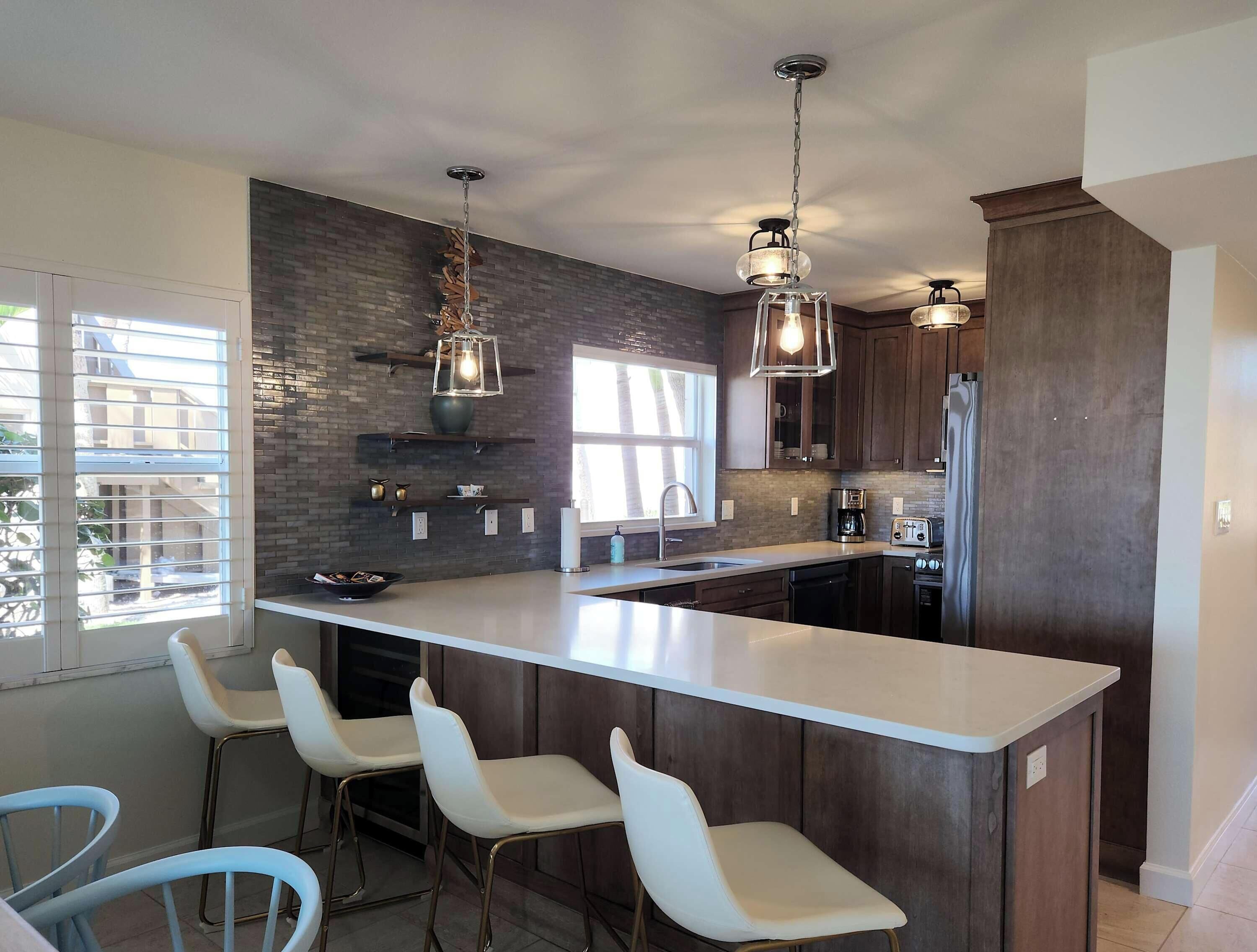 A modern kitchen with a large island, bar stools, pendant lighting, and a brick backsplash.