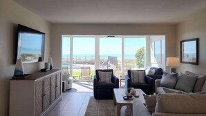 A spacious living room with a large sliding glass door leading to a balcony with a view of the sea. There are two sofas, a coffee table, and a TV on a cabinet. - Whitney Beach 321 2brm beachfront (Longboat Key)