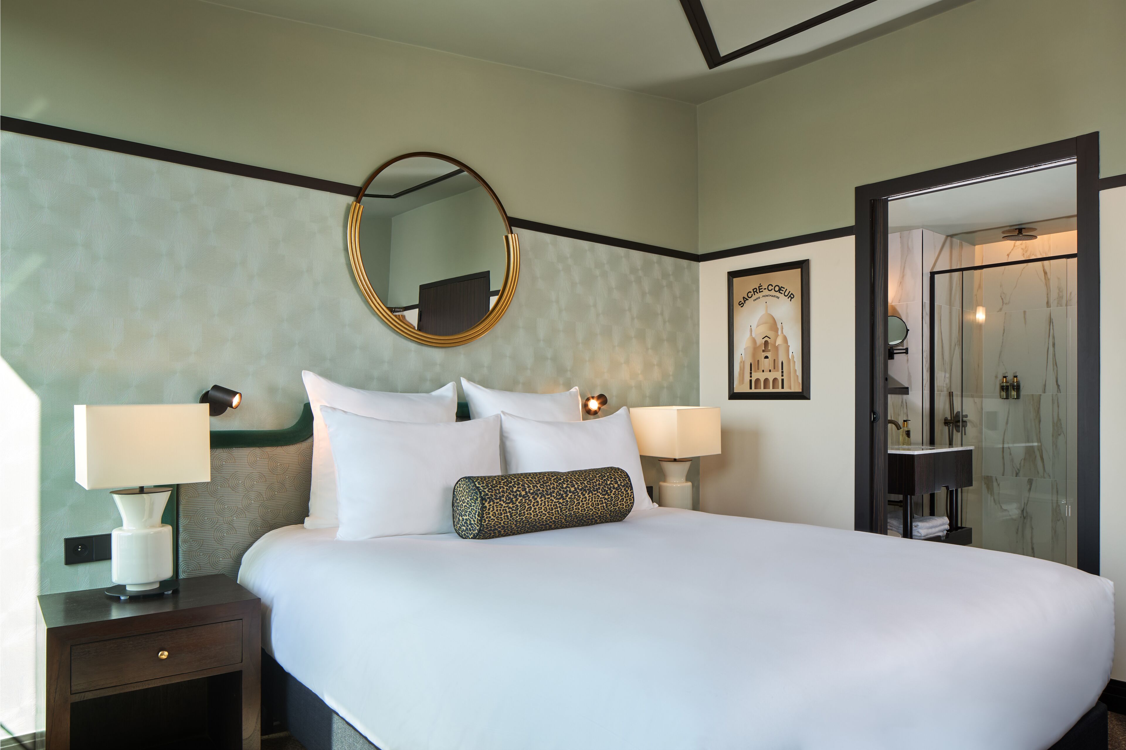 Una habitación de hotel con una cama grande, dos lámparas de noche, una mesita de noche, un espejo redondo, un cuadro enmarcado y una zona de ducha con mampara de vidrio.