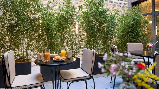 An outdoor seating area with a stone wall, potted plants, and a table set for two with a croissant and a glass of orange juice. - Maison Cassandre (Paris)