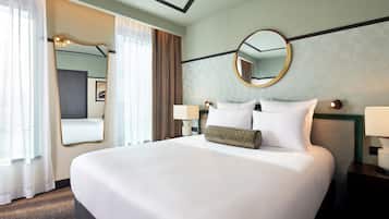 Habitación de hotel con una cama grande, dos mesitas de noche con lámparas, un espejo redondo y una ventana con cortinas.