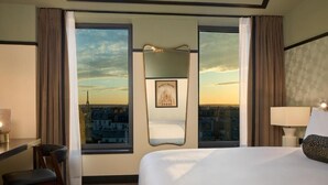 A hotel room with a large bed, a desk, and a chair. There is a large window with a view of a cityscape and a framed picture on the wall. - Maison Cassandre (Paris)