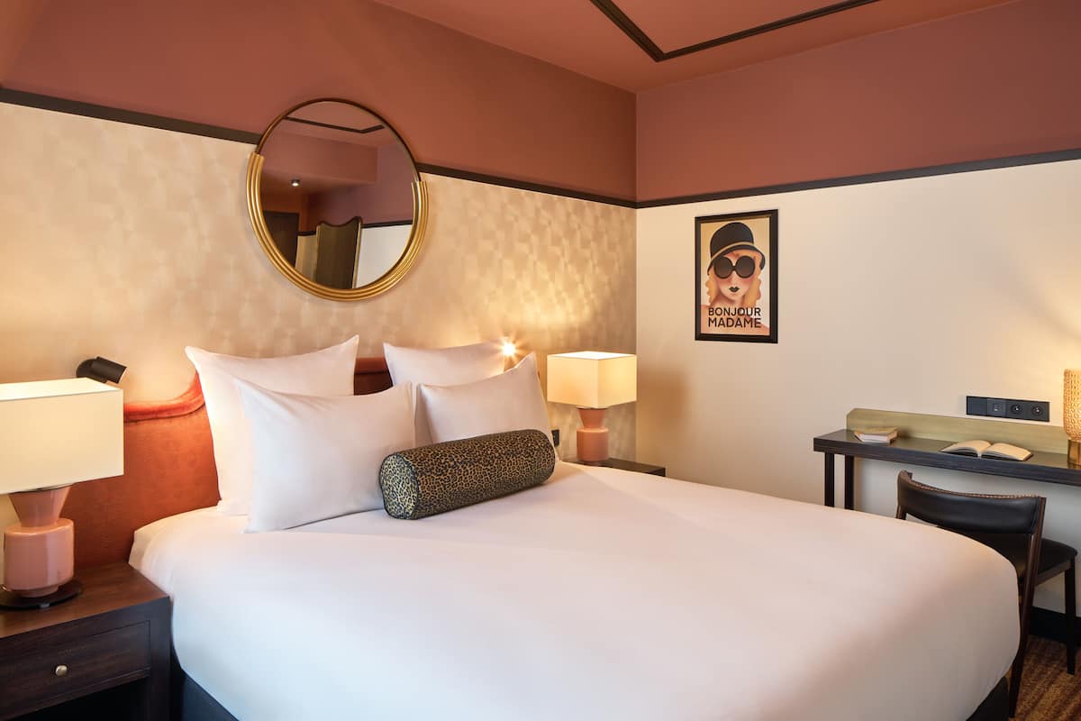 Un cuarto de hotel con una cama grande, dos mesitas de noche con lámparas, un escritorio con silla, un espejo redondo y un cartel en la pared.