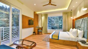A hotel room with a large bed, a desk, a chair, and a balcony with a view of trees.