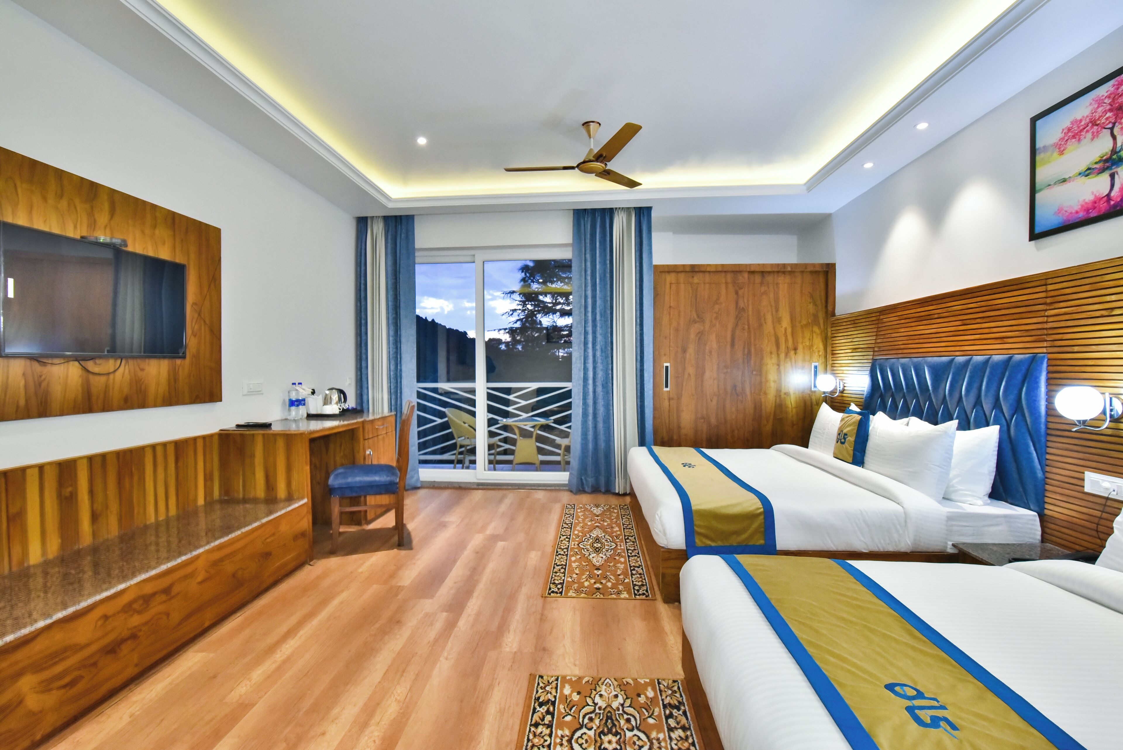 Une chambre d’hôtel avec un plancher en bois, un téléviseur fixé au mur, un lit recouvert d’une courtepointe bleue et jaune, un bureau avec une chaise et un balcon donnant sur des arbres.