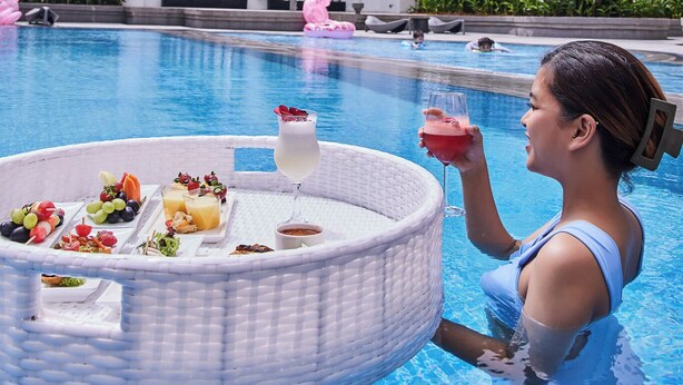 Ein Poolbereich mit Esstisch und Getränken, umgeben von Palmen und einem modernen Gebäude im Hintergrund.