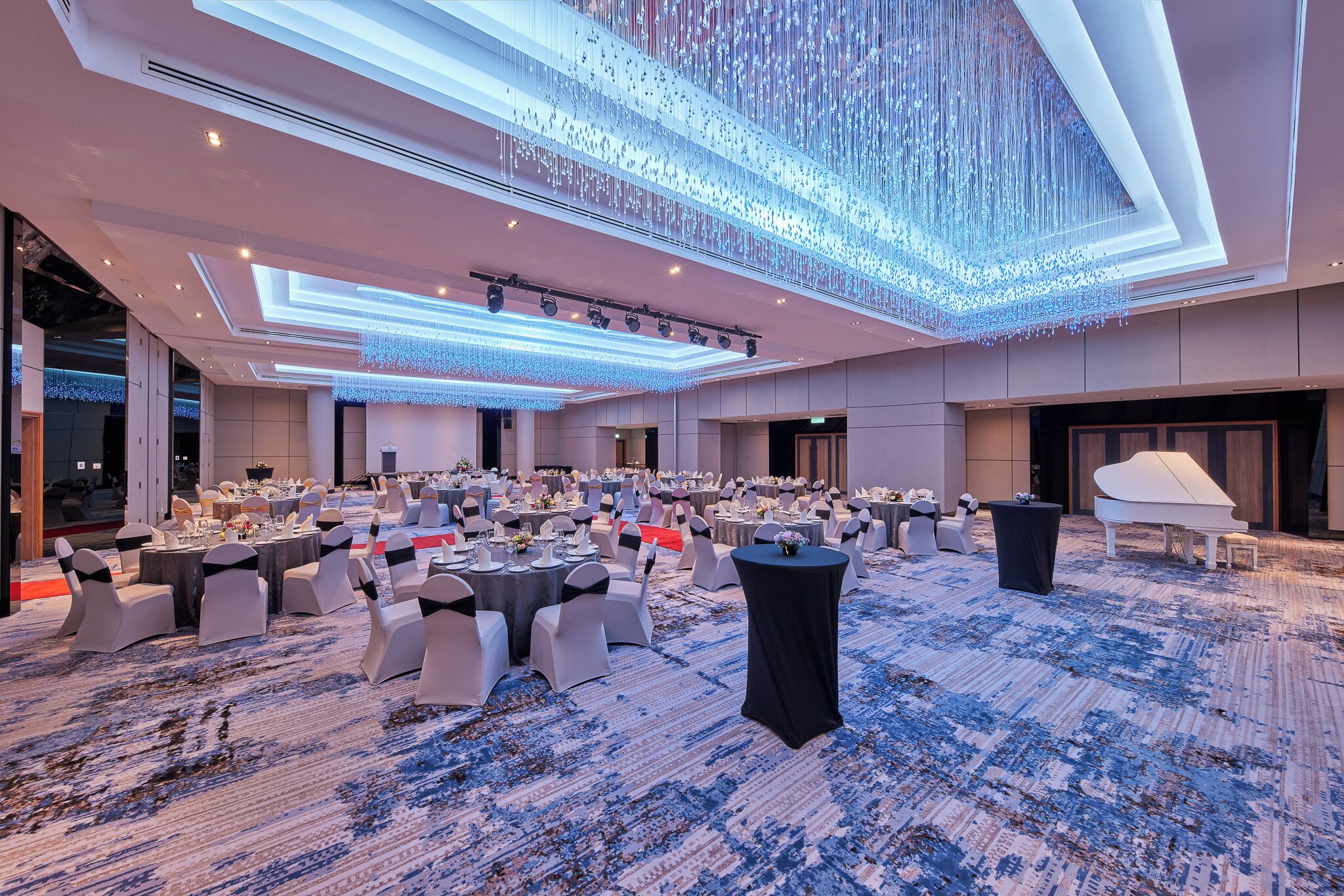 A spacious banquet hall with round tables and chairs, a grand piano, and a modern chandelier.