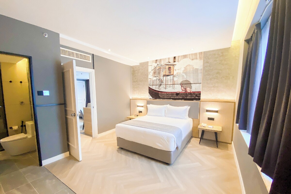 A modern hotel room with a large bed, bedside tables, a bathroom with a toilet and sink, and a painting on the wall.