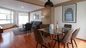 A spacious living area with a glass dining table, comfortable seating, a sofa, and a large window.