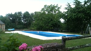 Ein Schwimmbecken, umgeben von einer Steinmauer, mit einem Holzzaun im Vordergrund und üppigem Grün im Hintergrund.